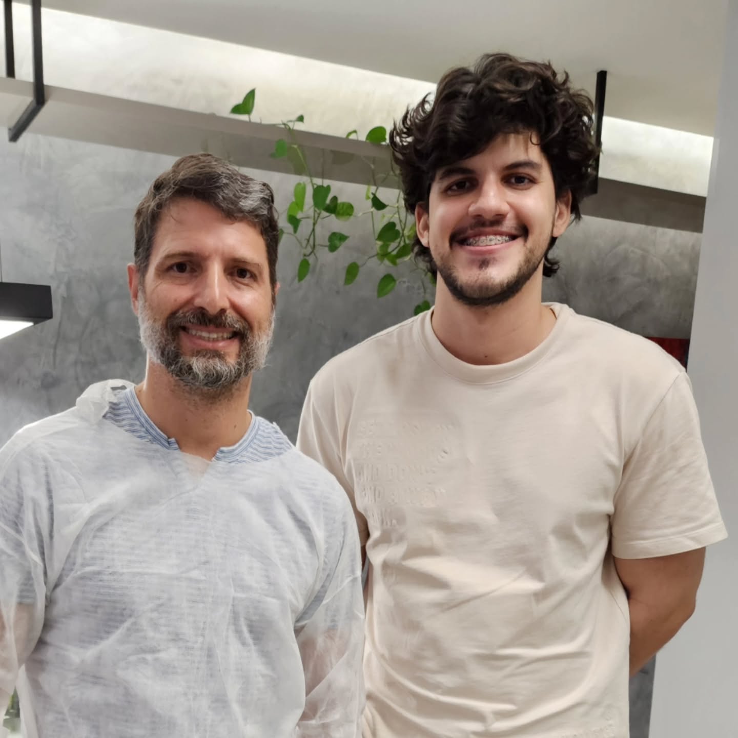 O Dr. Luiz Fernando, de avental descartável, ao lado do paciente, que sorri ainda com o aparelho